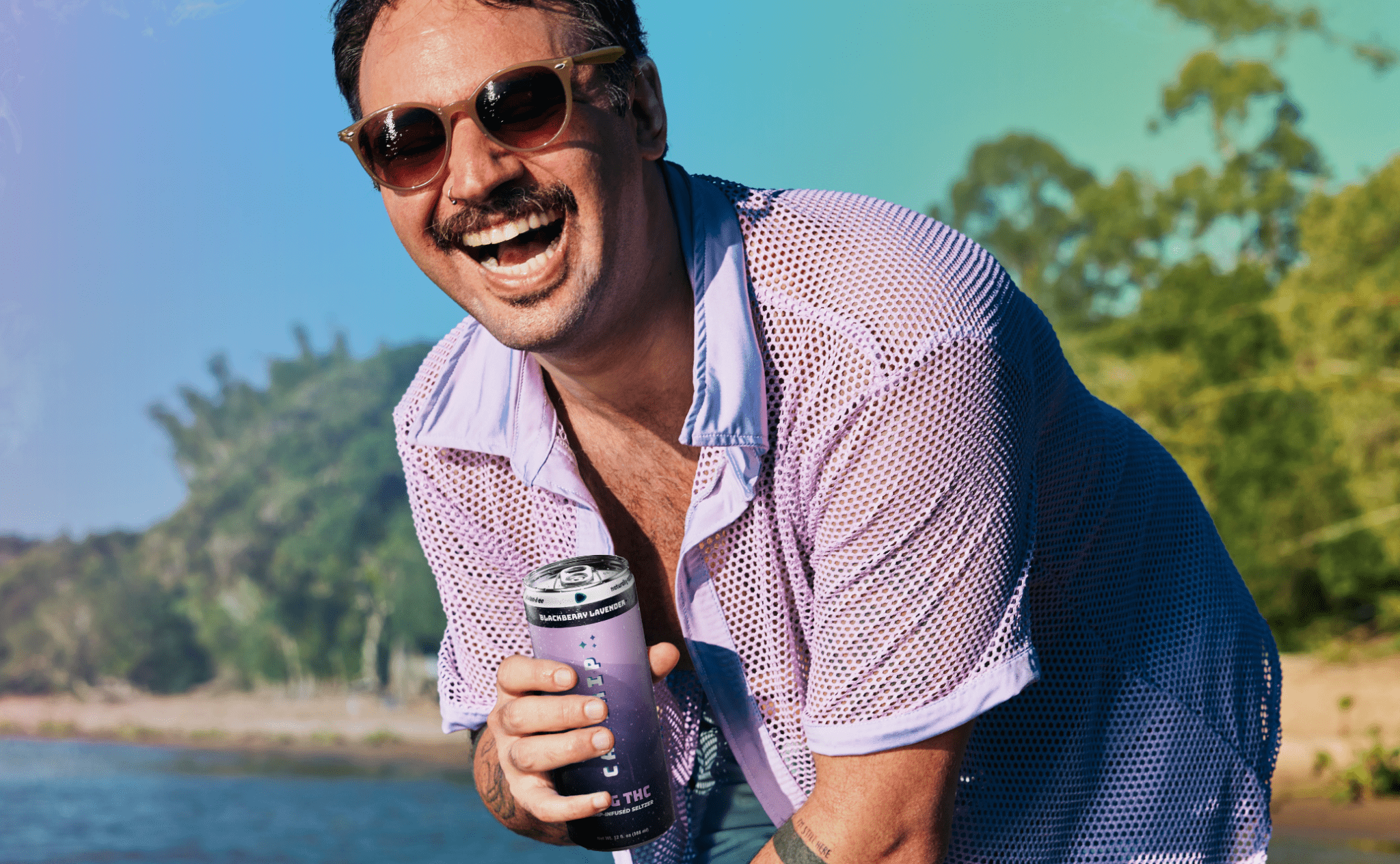 A mean wearing sunglasses and a lavender shirt laughs on the beach with a can of Blackberry Lavender THC Seltzer in his hand.