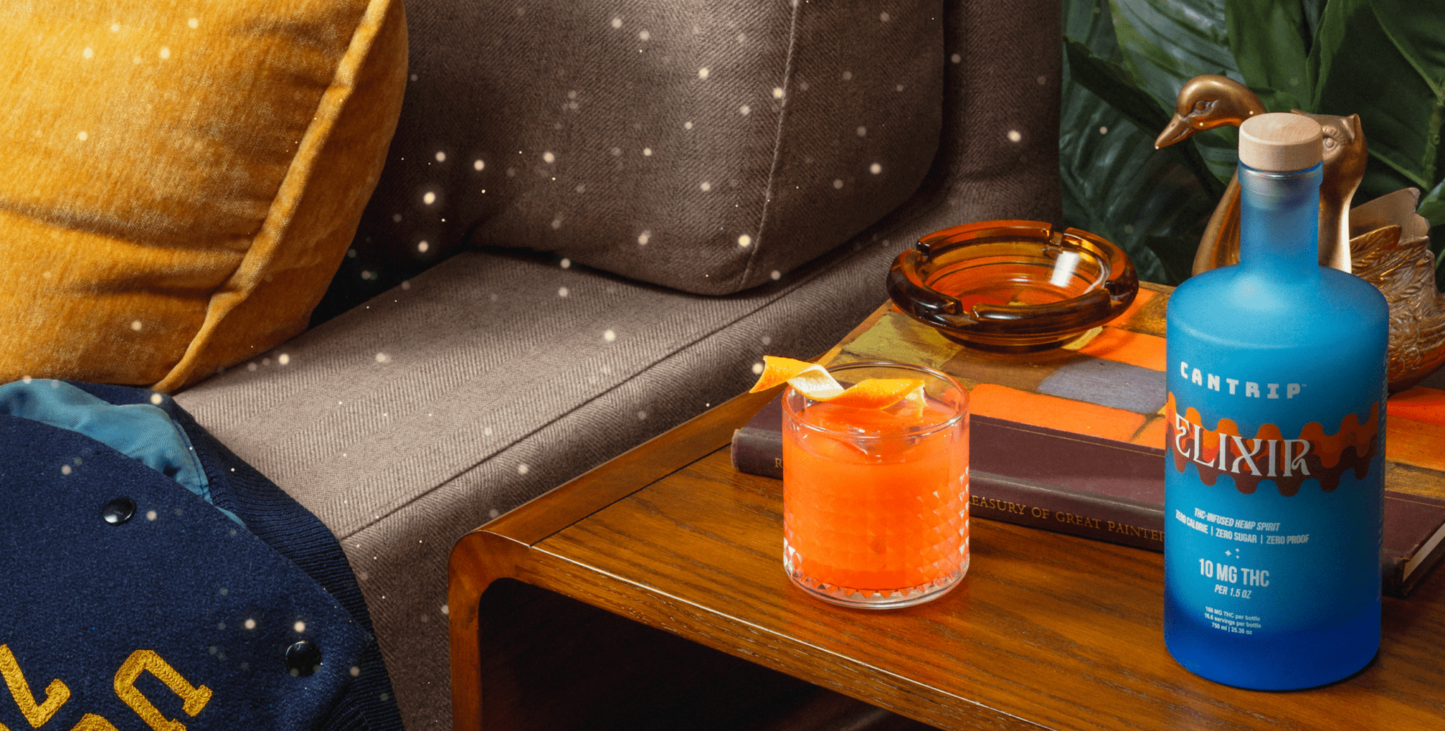 A blue bottle of zero-proof Cantrip Elixir and a non-alcoholic cocktail with a twist of orange sit on an end table.