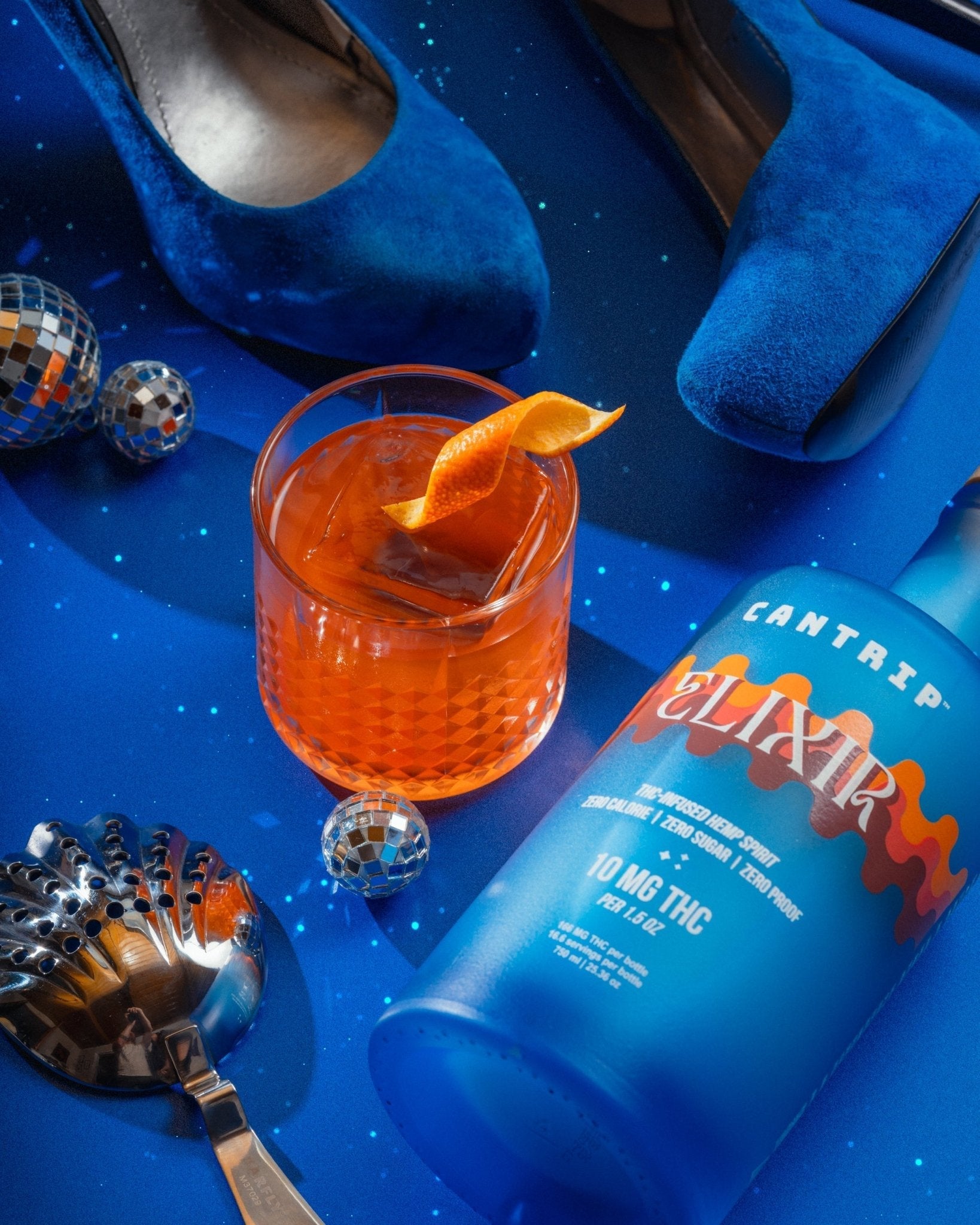 A blue bottle of Cantrip Elixir lying next to an orange-colored Negroni on a blue surface. They're surrounded by little disco balls, blue velvet shoes, and a silver spoon.