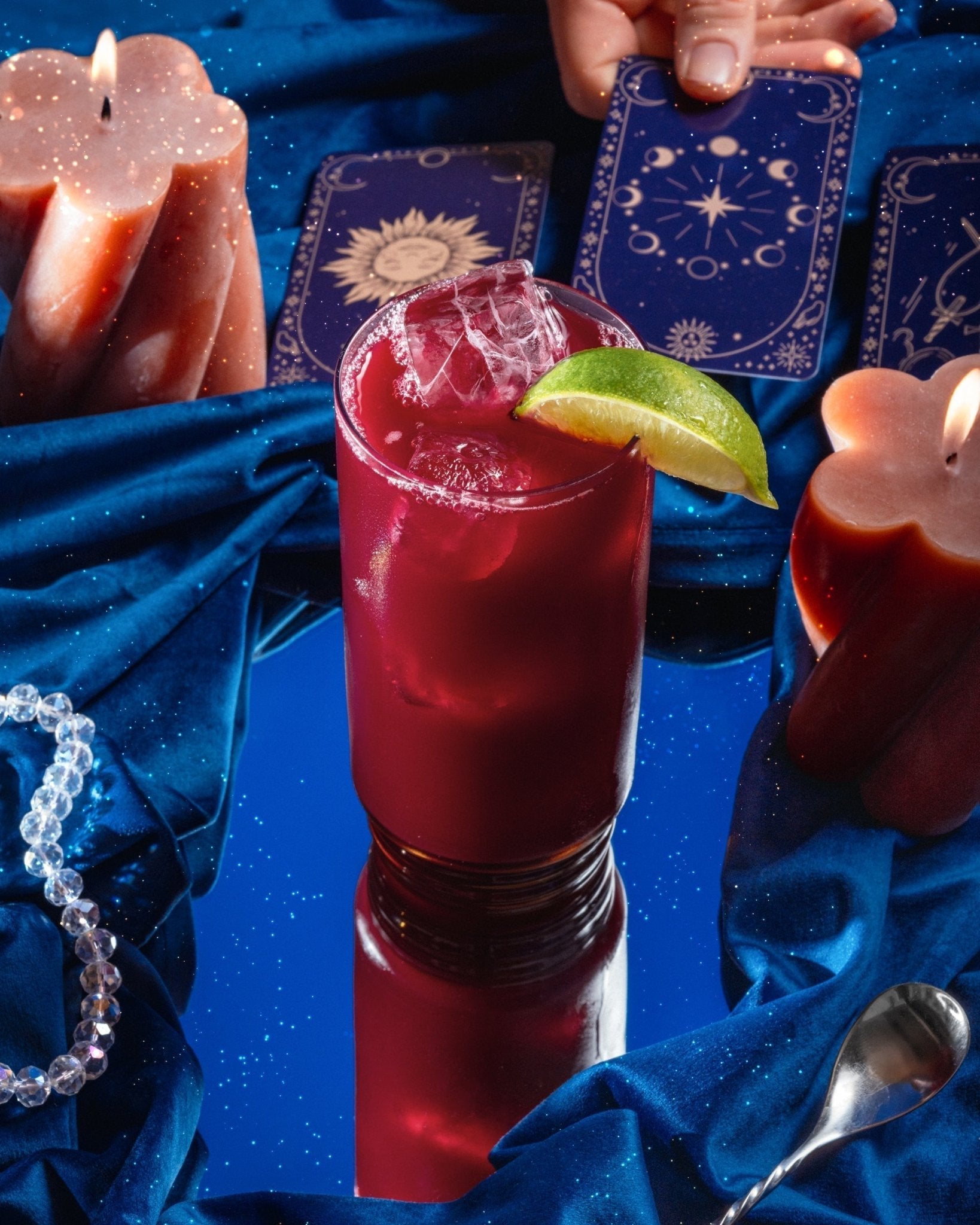 A red, non-alcoholic Elixir on the Cape cocktail garnished with a lime wedge and surrounded by candles as someone plays with tarot cards