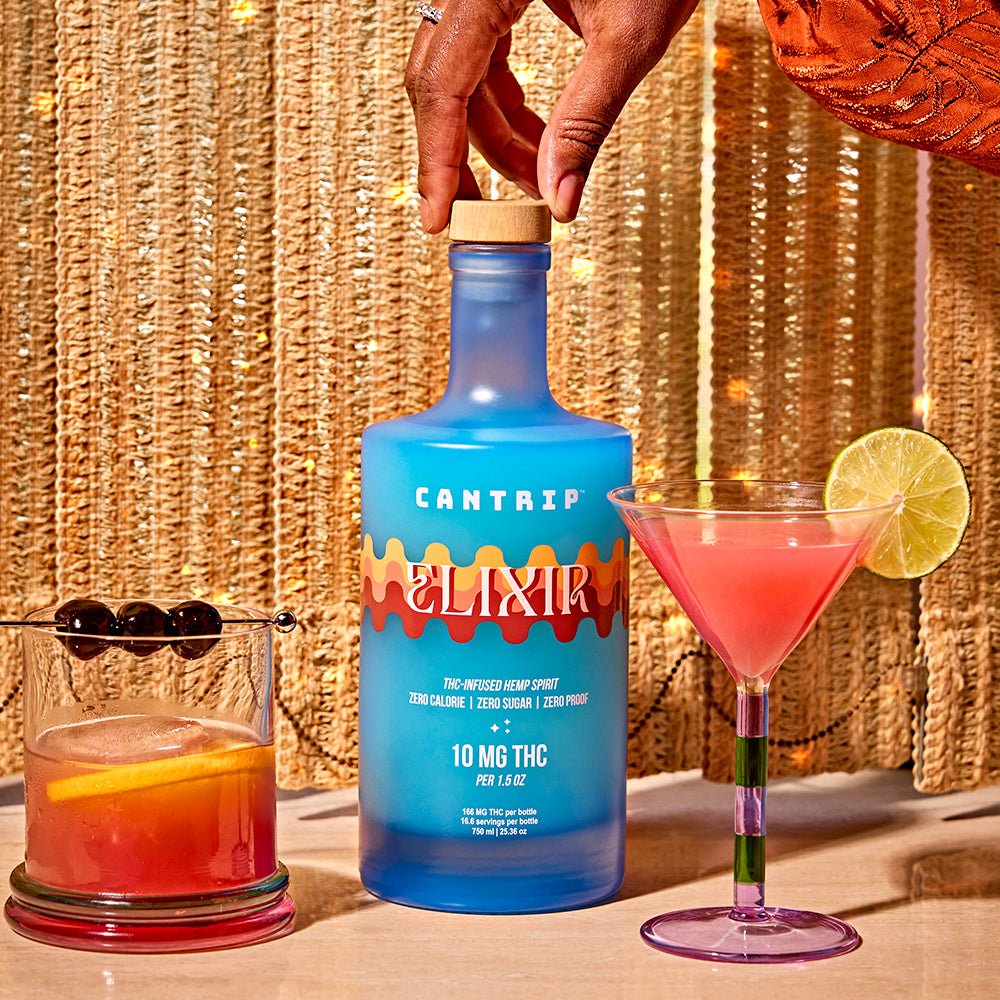 A hand reaching in to close a blue bottle of Cantrip Elixir; the bottle is set between an old fashioned and paloma, both made with the THC infused spirit.