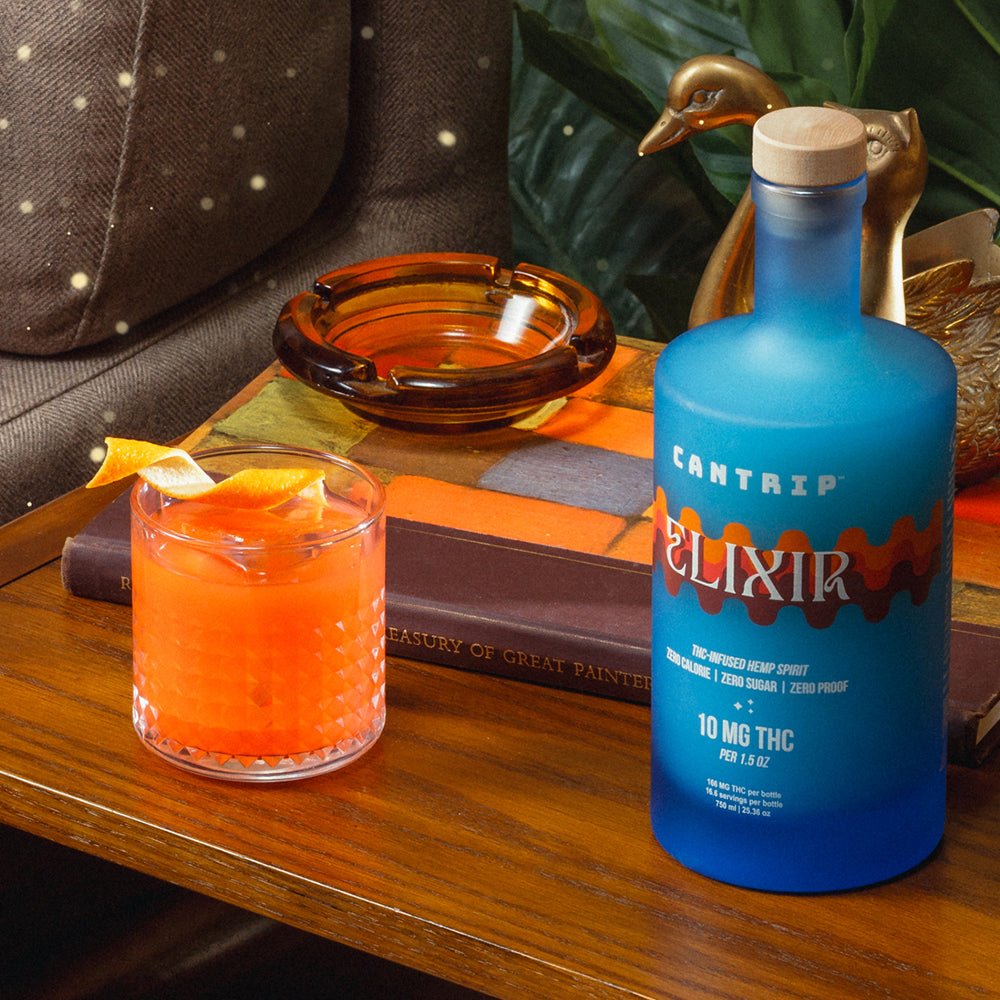 A blue bottle of Cantrip Elixir and a non-alcoholic cocktail with a twist of orange sit on an end table.