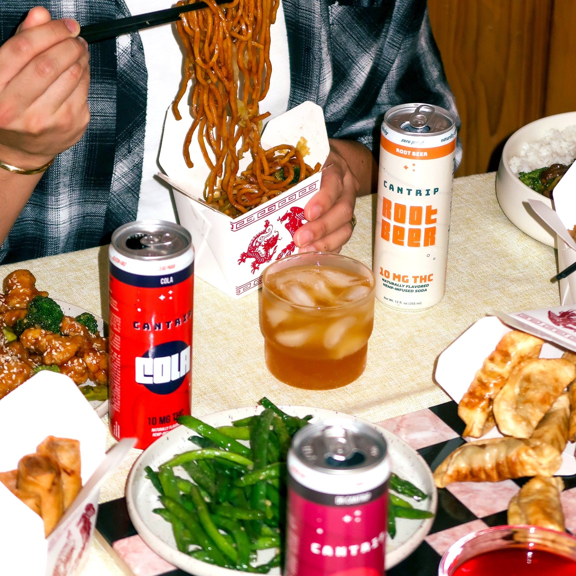 Someone in a plaid shirt eating Dan Dan noodles with chopsticks; Chinese takeout and Cantrip THC sodas are on the table.