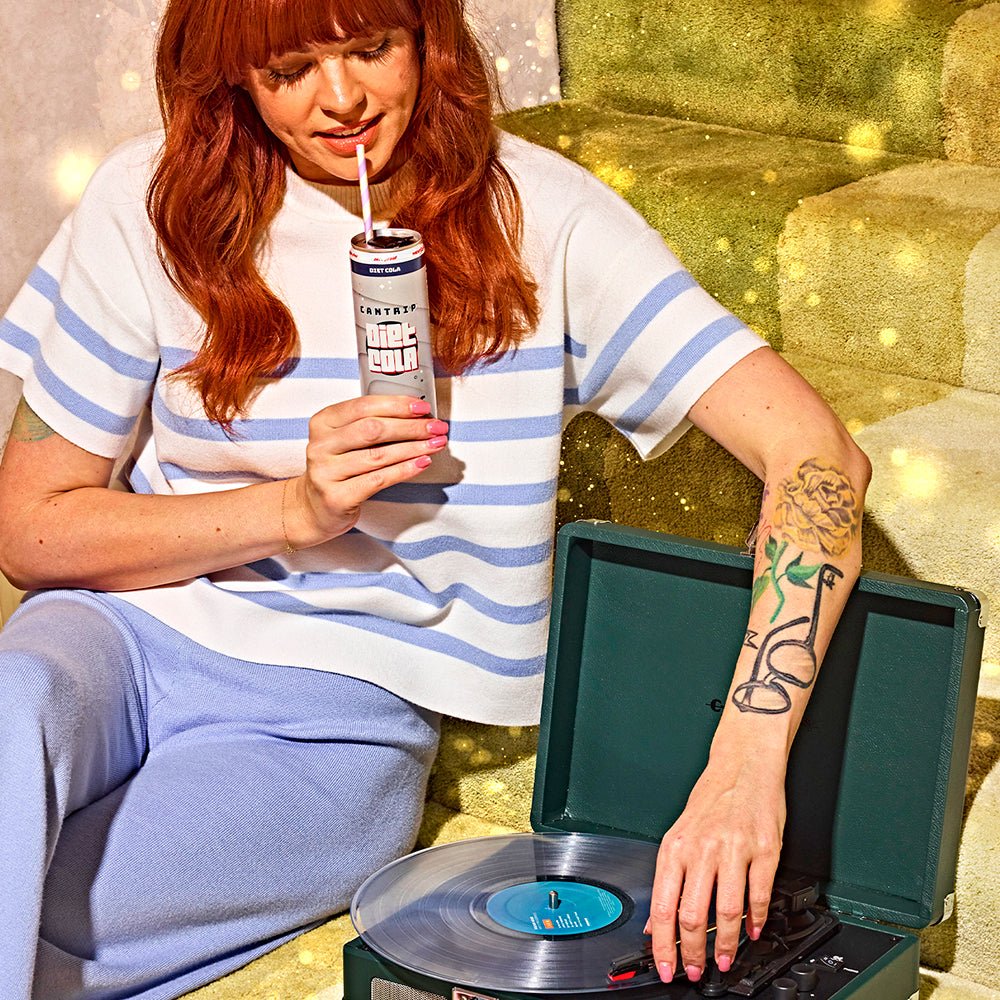 A woman with red hair and tattoos, holds a can of Cantrip THC Diet Cola, while placing the needle on a vinyl record.
