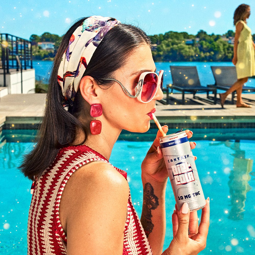 A woman wearing big red mirrored sunglasses, a red and white crocheted top, and red earrings sips a Cantrip Diet Cola through a straw while standing poolside.