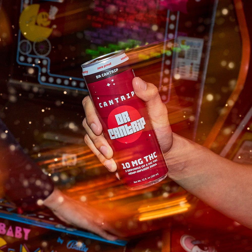 Hand holding a red can labeled Dr Cantrip with a blurred background in an arcade.
