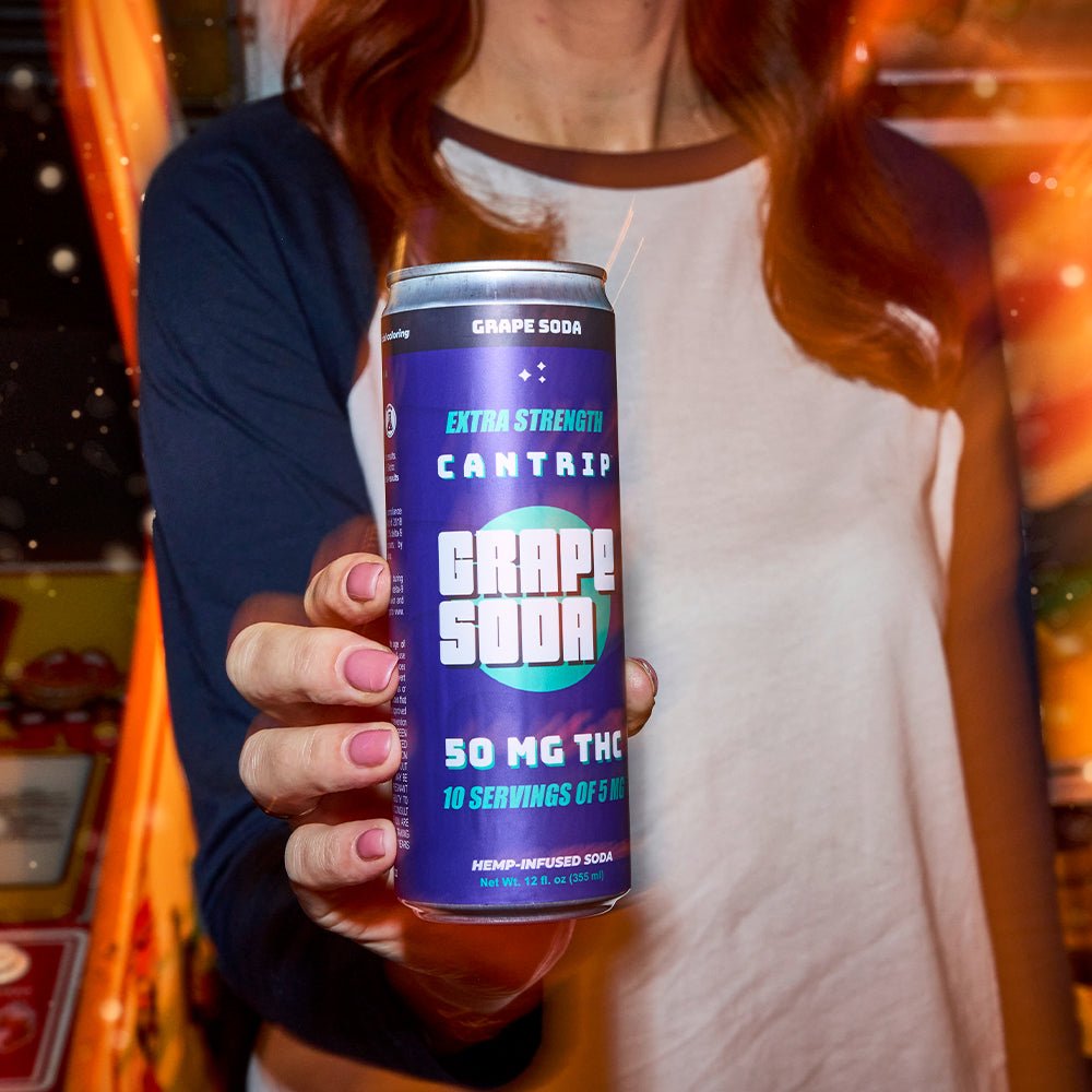 A manicured hand with lilac nail polish reaching toward the camera with a purple can of 50 mg Cantrip Grape Soda