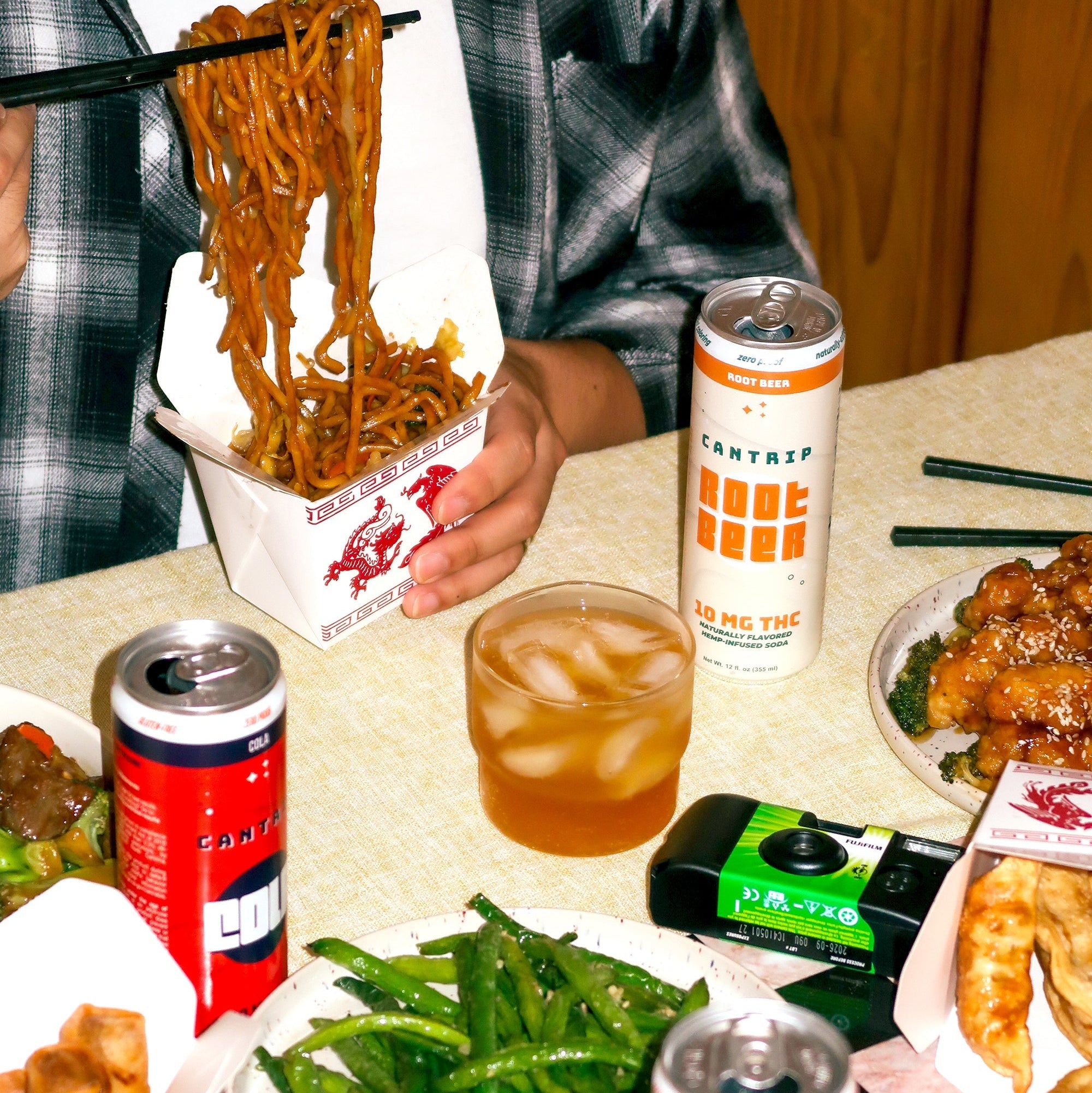 Someone in a plaid shirt eating Dan Dan noodles with chopsticks; Chinese takeout, Cantrip THC sodas , and a disposable camera are on the table.