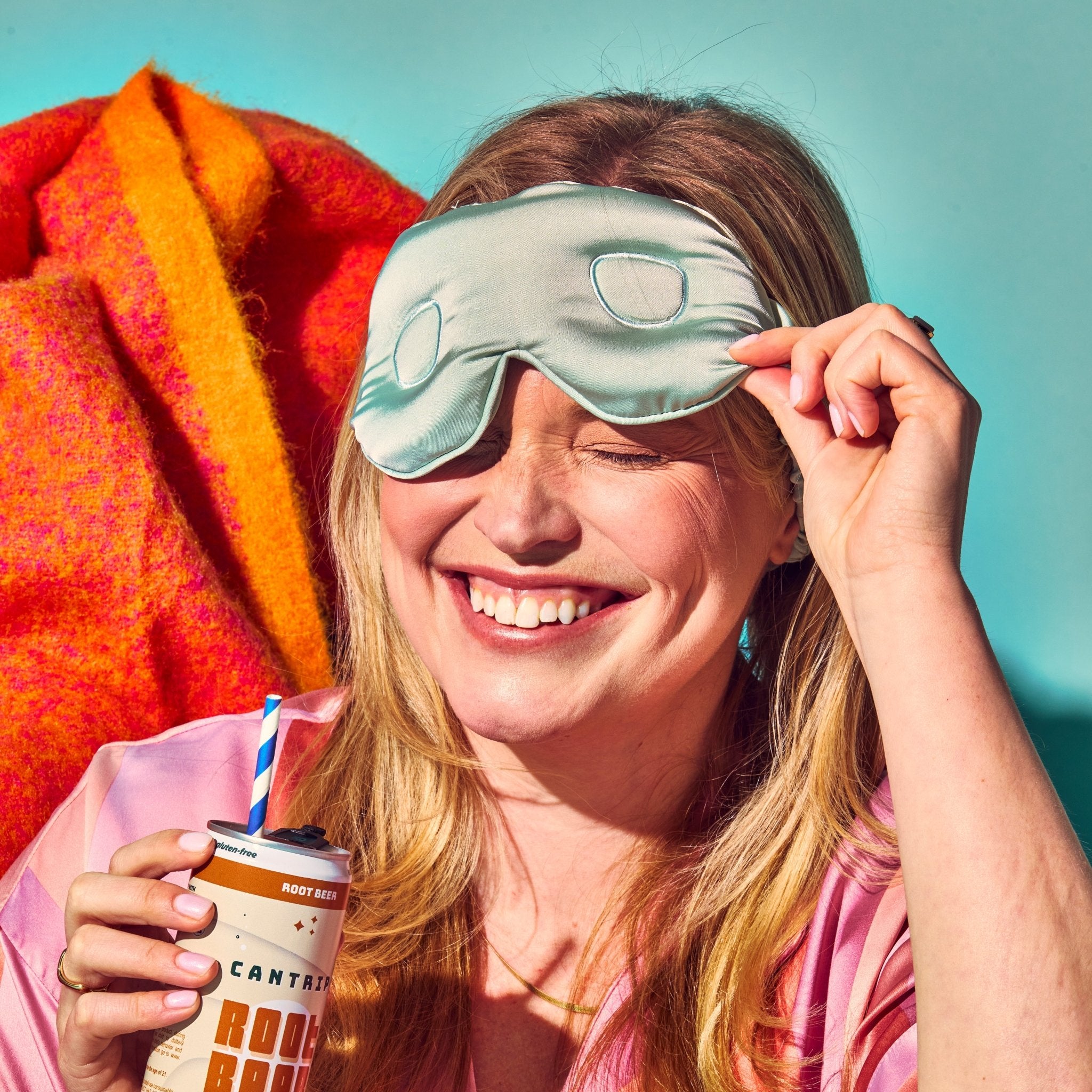 A smiling woman with blonde hair is pulling back her sleeping mask. She's holding a can of 50 MG Cantrip THC root beer.