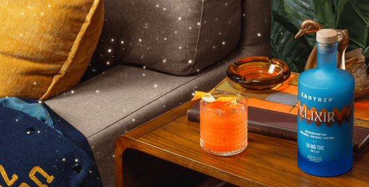 A blue bottle of zero-proof Cantrip Elixir and a non-alcoholic cocktail with a twist of orange sit on an end table.