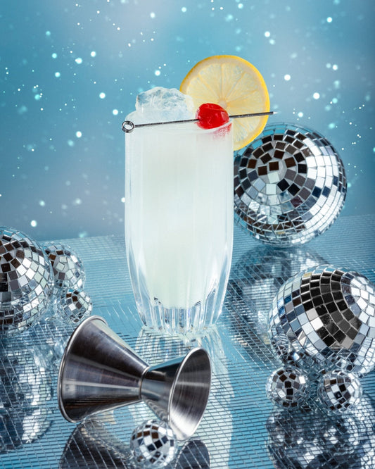 A zero-proof Elixir Collins in a tall glass, garnished with a lemon and cherry, and surrounded by mirrored balls and a jigger