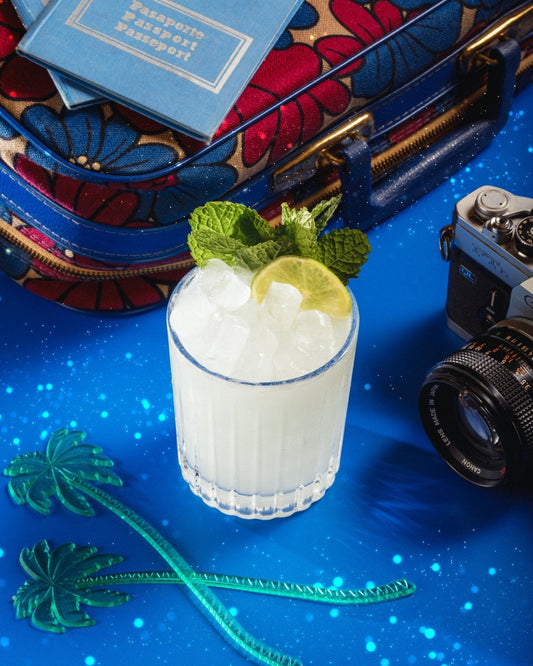 A non-alcoholic Mai Tai cocktail, made with Cantrip Elixir and garnished with a lemon wedge and mint, on blue surface with a camera, passport, and colorful suitcase in the background.