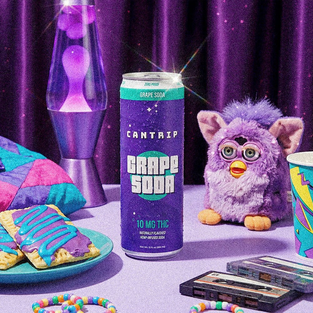 Cantrip Grape Soda THC can on a table with a lava lamp, cassette tapes, frosted pastries, and a plush toy against a purple backdrop